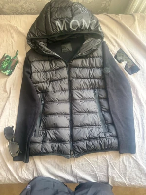 Moncler cardigan - Snygg Down Hybrid Jacket Black med quiltad front och stickade ärmar. Jackan har en stor huva med textdetalj och två dragkedjeförsedda fickor framtill. Perfekt för kalla vinterdagar och ger en modern look. Det svårt att ta bild på taggarna men om man kollar några kan man se den där med är det moncler cardigan. Priset kan diskuteras.