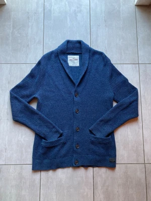 Blå kofta från Hollister - Mysig blå kofta från Hollister med grovstickad struktur och sjalkrage. Koftan har fem knappar framtill, två fickor och ribbade muddar vid ärmslut och nederkant. Perfekt för lager på lager och chill stil. Strl:L
