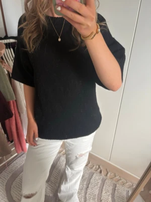 Svart stickad tröja med kort ärm - En svart stickad tröja med korta, vida ärmar och ribbade muddar vid ärmslut och nederkant. Tröjan har en rak passform och rund halsringning. Perfekt att styla med jeans eller kjol för en enkel och snygg look.