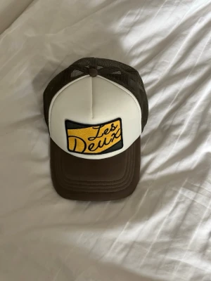 Brun och vit truckerkeps Les Deux - Snygg truckerkeps från Les Deux i brunt och vitt med gul och svart patch framtill. Kepsen har mesh på sidorna och bak för extra ventilation samt justerbar snapback. Perfekt för en avslappnad streetwear-look.