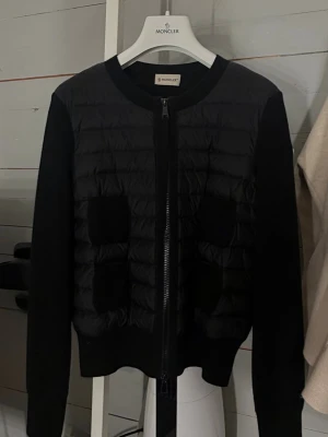Svart cardiganjacka från Moncler - Snygg svart cardiganjacka från Moncler med quiltad framsida och stickade ärmar. Jackan har rund halsringning, dragkedja framtill och två fickor. Perfekt för lager-på-lager och ger en stilren look. 