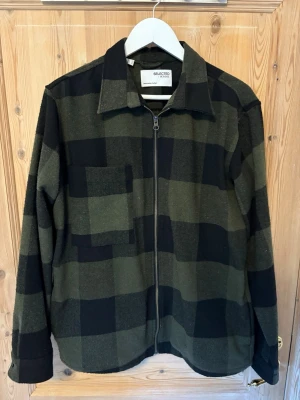 Grönrutad overshirt från Selected Homme - Snygg overshirt i grönt och svart rutmönster från Selected Homme. Jackan har dragkedja framtill, klassisk krage och en bröstficka. Tillverkad i ett mjukt tyg som passar perfekt för lager på lager. Perfekt för dig som gillar en avslappnad men stilren look.