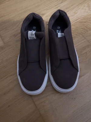 Bruna slip-on sneakers med vit sula - Säljer dessa skor oanvända helt nya Stilrena bruna slip-on sneakers med chunky  vit sula. Ovandelen är i textil och skon har en minimalistisk design utan snörning, vilket ger en clean och modern look. Perfekta för dig som gillar enkelhet och komfort i vardagen.