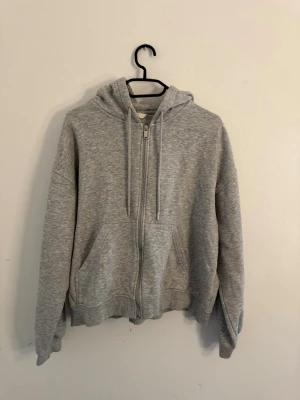 Grå zip-hoodie med fickor - En klassisk grå hoodie med dragkedja framtill och två stora fickor. Tröjan har huva med dragsko och ribbade muddar vid ärmslut och nederkant. Perfekt för en avslappnad och stilren look.