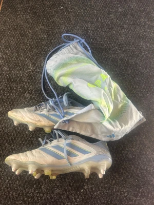 Ljusblå Adidas Copa fotbollsskor - Säljer ett par ljusblå Adidas Copa fotbollsskor med klassiska tre ränder i blått på sidan. Skorna har snörning och dobbar i metall och plast för bra grepp på planen. Medföljer en matchande blå och grön skopåse. Perfekta för dig som vill ha stilrena och funktionella skor. Skorna är lite slitna , dom är elite och äkta från unisport. Pris kan diskuteras via snabb affär!