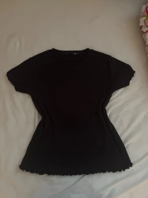 Svart top - En svart ribbad topp med korta ärmar och volangkant vid ärmslut och nederkant. Toppen har en figurnära passform och är tillverkad i ett mjukt stretchigt material som känns bekvämt mot huden. Perfekt att styla med jeans eller kjol.