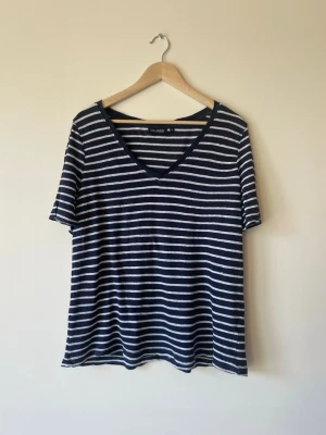 Randig marinblå t-shirt med v-ringning - Snygg marinblå t-shirt med vita horisontella ränder och v-ringad hals. T-shirten har korta ärmar och en avslappnad passform, perfekt för en chill och stilren look. Tillverkad i mjukt material som känns skönt mot huden.