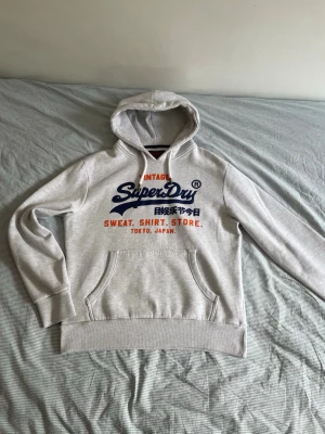 Grå Superdry hoodie med tryck - Snygg ljusgrå hoodie från Superdry med stor logga och japansk text på bröstet i blått och orange. Klassisk känguruficka framtill och mjuk insida. Perfekt för dig som gillar streetwear och vill ha något bekvämt och stilrent.