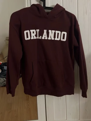 Vinröd hoodie med ORLANDO-tryck - Säljer en vinröd hoodie med vit text 'ORLANDO' på bröstet. Hoodien har en klassisk huva och en stor magficka framtill. Tillverkad i mjukt och skönt material, perfekt för chill dagar. Märkt med storlek 150.