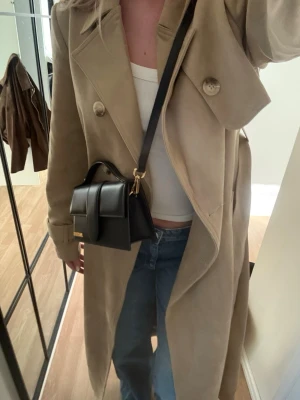 Beige lång trenchcoat med bälte - Säljer denna fina nästan slutsålda beigea kappan ifrån Lovisa Worges kollektion med NAKD. Använd få tal gånger så är i väldigt bra skick! Köpt för 1399