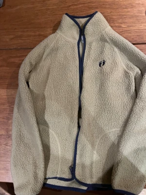 Beige fleecejacka  - Mysig beige fleecejacka med hel dragkedja och blå kantband. Jackan har hög krage och två fickor framtill. Perfekt för kyliga dagar och enkel att matcha med andra plagg.