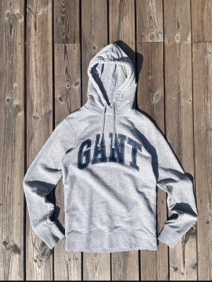 Grå hoodie från GANT med tryck - Snygg grå hoodie från GANT med stor logga i mörkblått på bröstet. Klassisk modell med huva och dragsko samt ribbade muddar vid ärmar och nederkant. Perfekt för en avslappnad och sportig stil.