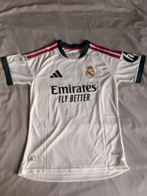 Real Madrid matchtröja Mbappé 10 - Säljer en vit Real Madrid matchtröja från Adidas med gröna detaljer och rosa ränder på axlarna. Tröjan har klubbmärke, sponsortryck och nummer 10 med Mbappé på ryggen. Tillverkad i lätt och ventilerande material, perfekt för fotbollsträning eller match.