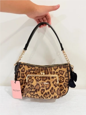 Leopardmönstrad handväska Juicy Couture - Leopardmönstrad handväska från Juicy Couture med guldfärgade detaljer och kedjehandtag. Väskan har en svart axelrem, en dekorativ rosett på sidan och en praktisk ytterficka med guldram. Perfekt statement piece för dig som gillar djurmönster.