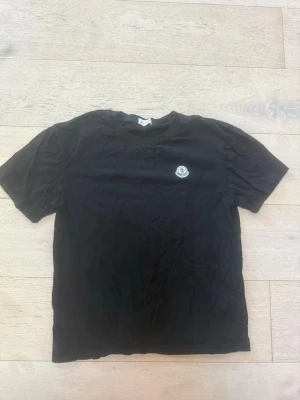 Svart t-shirt med liten logga - En svart t-shirt i klassisk modell med rund hals och korta ärmar. På bröstet sitter en liten rund logga som ger en clean och stilren look. Perfekt till jeans eller shorts för en enkel och snygg outfit.