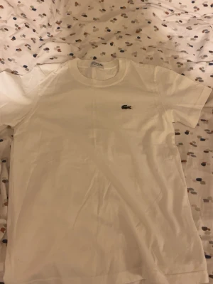 Vit t-shirt  - Klassisk vit t-shirt med den ikoniska broderade krokodilloggan på bröstet. T-shirten har rund hals och korta ärmar, perfekt för en clean och stilren look. Tillverkad i mjuk bomull för maximal komfort.