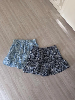 Två blommiga kortkjolar i blå toner - Två korta kjolar med blommigt mönster, en i ljusblått och en i mörkblått. Båda har innershorts, resår i midjan och är tillverkade i ett lätt, luftigt material som passar perfekt för varmare dagar.