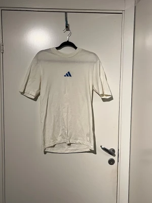 Vit t-shirt med blå Adidas-logga - Säljer en enkel och clean vit t-shirt från Adidas med den klassiska blå loggan på bröstet. T-shirten har korta ärmar och rund halsringning. Perfekt till jeans eller shorts för en avslappnad look.