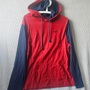 Röd och marinblå hoodie med huva - Snygg hoodie från Hollister i rött med marinblå ärmar och detaljer. Klassisk huva med dragsnören och stor magficka framtill. Liten Hollister-logga broderad på bröstet. Perfekt för en avslappnad och cool stil.