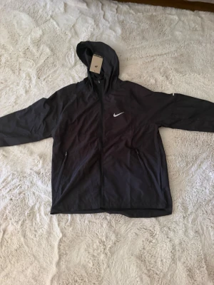 Svart vindjacka med huva från Nike - Snygg svart vindjacka från Nike med hel dragkedja, huva och två sidofickor med dragkedja. Jackan har en vit Nike-logga på bröstet och är tillverkad i lätt, vindtätt material. Perfekt för träning eller promenader när det blåser.