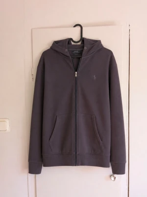Polo Ralph Lauren Zip Hoodie – Strl S 🐎 - Stilren full-zip hoodie från Polo Ralph Lauren i mörkgrå/charcoal färg. Klassisk modell med huva, dragkedja hela vägen framtill, kängurufickor och diskret broderad Polo-logga på bröstet. Storlek: S Färg: Mörkgrå / charcoal Skick: Bra begagnat skick med normalt bruksslitage – se bilder. Detaljer: Full zip, huva, två framfickor och broderad Ralph Lauren-logo. Väldigt lättmatchad hoodie som passar perfekt till vardags. Hör gärna av dig vid frågor eller om du vill ha mått! 📏