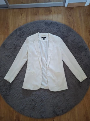 Vit kavaj från H&M, storlek 38 - Stilren vit kavaj från H&M i storlek 38. Klassisk modell med smal passform, långa ärmar och två fickor framtill. Tillverkad i ett lätt tyg som ger en snygg och fräsch look. Perfekt att matcha med jeans eller kjol för en trendig outfit.