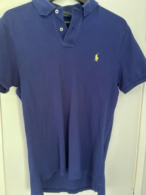 Blå pikétröja från Polo Ralph Lauren - Klassisk blå pikétröja från Polo Ralph Lauren med korta ärmar och gul broderad logga på bröstet. Tröjan har krage, knappar vid halsen och är tillverkad i bomull. Perfekt för en stilren och avslappnad look.