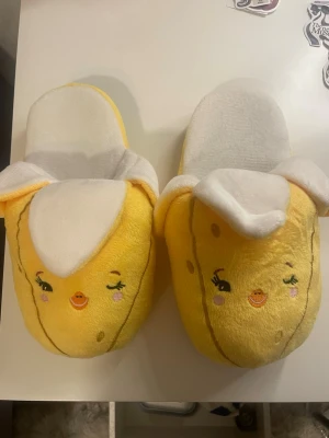 Gula banan-tofflor med ansikte - Roliga och mysiga tofflor i form av bananer med broderade ansikten och rosa kinder. Tofflorna är gula med vita detaljer som liknar bananskal och är gjorda i mjukt plyschmaterial. Perfekta för dig som vill ha något unikt och bekvämt hemma.