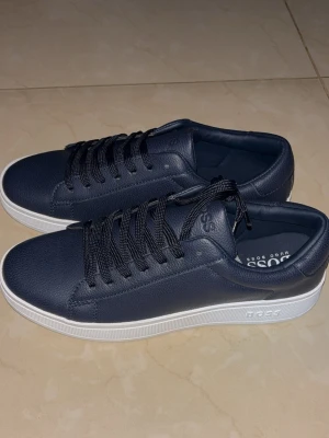 Mörkblå sneakers från BOSS - Stilrena mörkblå sneakers från BOSS med ovandel i slätt läder och vita, tjocka sulor. Klassisk låg modell med svarta skosnören och diskret BOSS-logga på sidan och plösen. Perfekta för en clean och modern look.