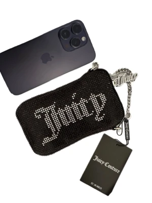 Mobilväska / aftonväska i strass. Juicy Couture - Helt ny & oanvänd svart axelväska / aftonväska / mobilväska från Juicy Couture med glittriga stenar och stor Juicy-logga i silver på framsidan.  Väskan har en silverfärgad kedja som axelrem och en snygg dragkedja med Juicy-detalj.  •Min iPhone 14 Pro med skal fick perfekt plats i väskan• Cirka mått i cm: Höjd: 16-17 Bredd: 10 ( den bredaste sidan) Djup: 3. 