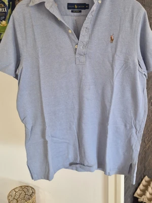 Ljusblå pikétröja från Ralph Lauren - Säljer en ljusblå pikétröja från Ralph Lauren med klassisk krage, korta ärmar och knappar vid halsen. Tröjan har den ikoniska broderade polospelaren på bröstet och är gjord i ett mjukt bomullsmaterial. Perfekt för en clean och stilren look.