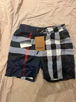 Burberry badbyxor - Snygga badbyxor från Burberry med klassiskt rutmönster i marinblått, vitt, svart och rött. De har resår i midjan för skön passform och är tillverkade i ett snabbtorkande material. Perfekta för stranden eller poolen.