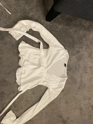 Vit omlottblus med knytband - Säljer en vit omlottblus med långa ärmar och knytband i midjan. Blusen har en lätt glansig yta och v-ringning framtill. Perfekt att styla med jeans eller kjol.