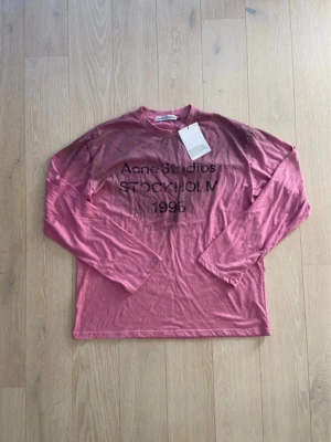 Rosa långärmad topp Acne Studios - Säljer en rosa långärmad topp från Acne Studios med tryck 'STOCKHOLM 1996' framtill. Toppen har rund halsringning och är gjord i mjuk bomull. Perfekt för dig som gillar streetwear och vill ha något stilrent men ändå unikt.