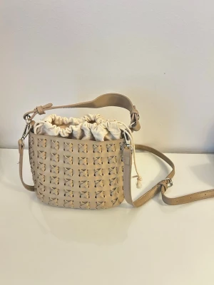 Beige flätad handväska med axelrem - Snygg beige handväska med flätad design och justerbar axelrem. Väskan har en tygpåse med dragsko inuti som stängning. modellen är bucket bag-inspirerad med trendig look.