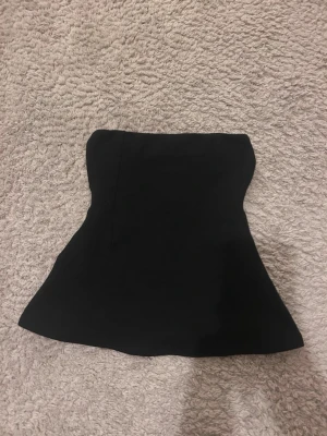 Svart tubtopp från Gina Tricot - Säljer en svart tubtopp från Gina Tricot i storlek XXS. Toppen är axelbandslös och har en enkel, stilren design som sitter tight runt överkroppen. Perfekt att matcha med höga jeans eller kjol för en trendig look.
