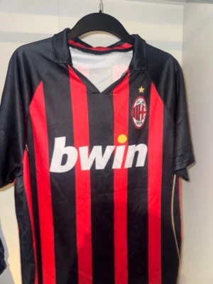 AC Milan matchtröja röd/svart - Säljer en klassisk AC Milan matchtröja i rött och svart med vertikala ränder. Tröjan har klubbmärke och Bwin-logga på bröstet samt guldfärgad stjärna ovanför loggan. 