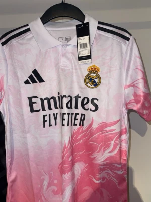 Real Madrid rosa matchtröja Adidas - Säljer en Real Madrid matchtröja från Adidas i vitt och rosa med drakmönster. Tröjan har korta ärmar, svart logga och klubbmärke på bröstet samt svart text 'Emirates Fly Better' framtill. Klassisk pikékrage och svarta ränder på axlarna.