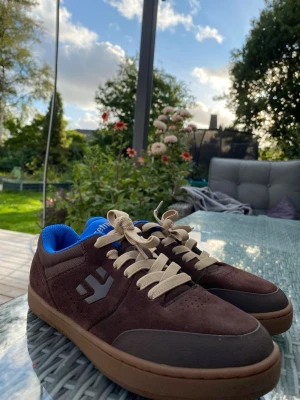 Bruna sneakers med blå insida - Säljer ett par bruna sneakers i storlek 36 med beigea skosnören och blå insida. Skorna har en låg profil och en gummisula i brunt. Ovandelen är i mockaliknande material med grå detaljer och en logotyp på sidan. Perfekta för dig som gillar stilrena och bekväma skor och även är dessa aldrig någonsin använda och jag köpte dessa för 350kr