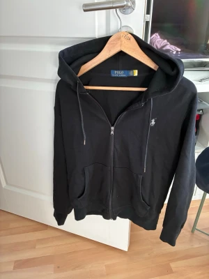 Svart hoodie från Polo Ralph Lauren - Svart hoodie med dragkedja från Polo Ralph Lauren. Klassisk design med två fickor framtill, justerbar huva med snören och diskret logga broderad på bröstet. Perfekt för en avslappnad och stilren look.