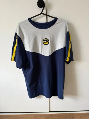 Nike blå och vit t-shirt med logga - Snygg t-shirt från Nike i marinblått och vitt med gul detalj på ärmarna och en cool Nike-logga framtill. T-shirten har rund hals och korta ärmar, perfekt för en sportig och avslappnad stil.