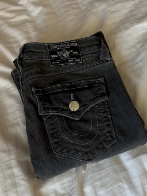 Svarta bootcut jeans True Religion - Säljer ett par svarta/gråa bootcut jeans från True Religion med de ikoniska bakfickorna. Jeansen har medium/låg midja. Perfekta för dig som gillar en avslappnad men ändå trendig look. PRISET KAN DISKUTERAS!!