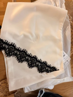 Zara mini kjol - Stilren vit kortkjol i satin med en dekorativ svart spetsdetalj som löper diagonalt över framsidan.  Byxkjol populära från Zara i Stl L 