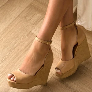 Beige kilklackade sandaler med rem - Snygga beige sandaler med hög kilklack och öppen tå. Skorna har en smal ankelrem med spänne och är tillverkade i ett material som liknar konstläder. Perfekta för dig som vill ha både höjd och komfort i sommarlooken.