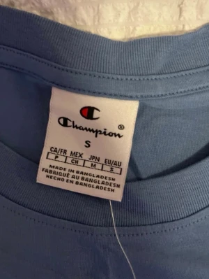 Blå Champion t-shirt i bomull - Snygg blå t-shirt från Champion med klassisk logga broderad på bröstet och liten logga på ärmen. Tillverkad i mjuk bomull och har en enkel, normal passform med rund hals och korta ärmar. Perfekt för en avslappnad och sportig stil.