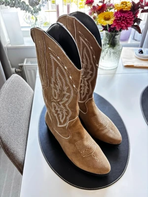 Beige cowboy boots i mocka - Snygga beige cowboy boots i mocka med klassiska vita broderier längs skaftet och framtill. Perfekta för en trendig look eller för en tema fest 🥳