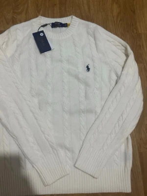 Vit kabelstickad tröja Polo Ralph Lauren - Kabelstickad vit tröja från Polo Ralph Lauren med rund halsringning och den klassiska broderade loggan på bröstet. Tröjan har ribbade muddar vid ärmslut och nederkant. Perfekt för dig som gillar stilrena och tidlösa plagg.