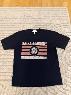 Mörkblå Saint Laurent t-shirt med tryck - Snygg mörkblå t-shirt från Saint Laurent med vitt och rosa tryck framtill. T-shirten har klassisk passform, rund hals och korta ärmar. Perfekt för dig som vill ha en stilren men ändå unik look.