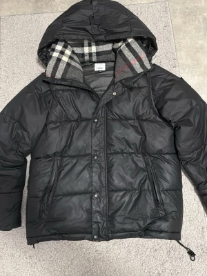 Svart puffer jacka / vest från Burberry M - Säljer en svart dunjacka från Burberry i storlek M. Jackan har huva och klassiskt rutigt foder i grått, vitt och rött. Två sidofickor med dragkedja och tryckknappar framtill. Perfekt för kalla vinterdagar och riktigt snygg design. Kan bli vest