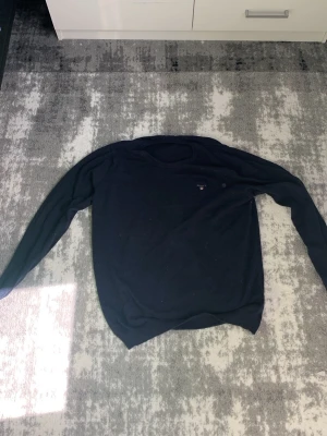 Mörkblå stickad tröja från GANT - Snygg mörkblå stickad tröja från GANT med rund halsringning och diskret logga broderad på bröstet. Tröjan har långa ärmar och ribbade muddar vid ärmslut och nederkant. Perfekt till en clean och klassisk stil.